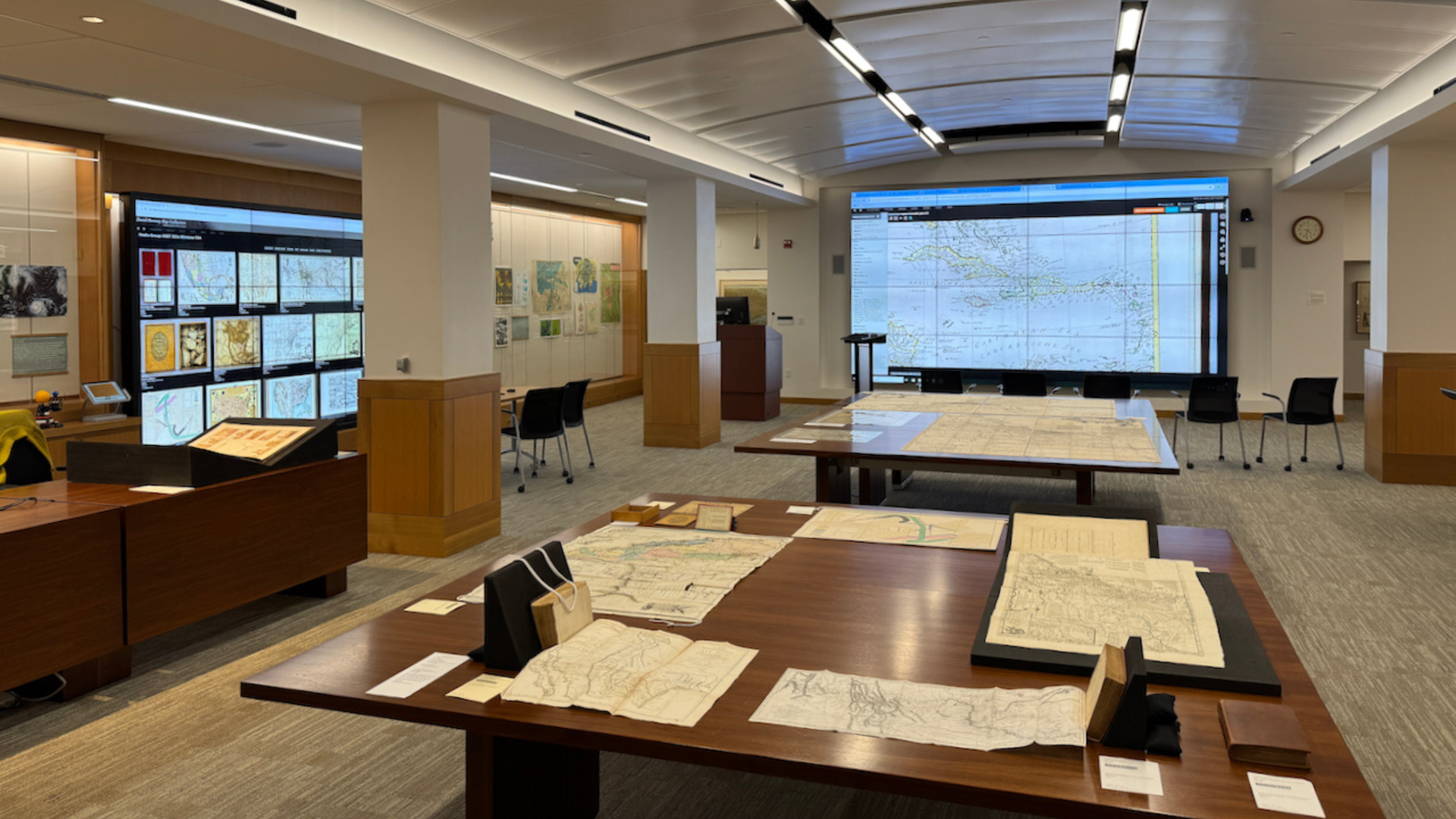 A room featuring large wallscreens showing digital images of maps with tables in the center displaying archival atlases and paper maps